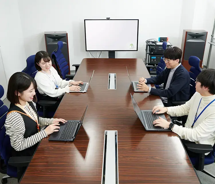 写真:会議テーブルで各自ノートPCを扱いながら打ち合わせをしている男性社員2名と女性社員2名