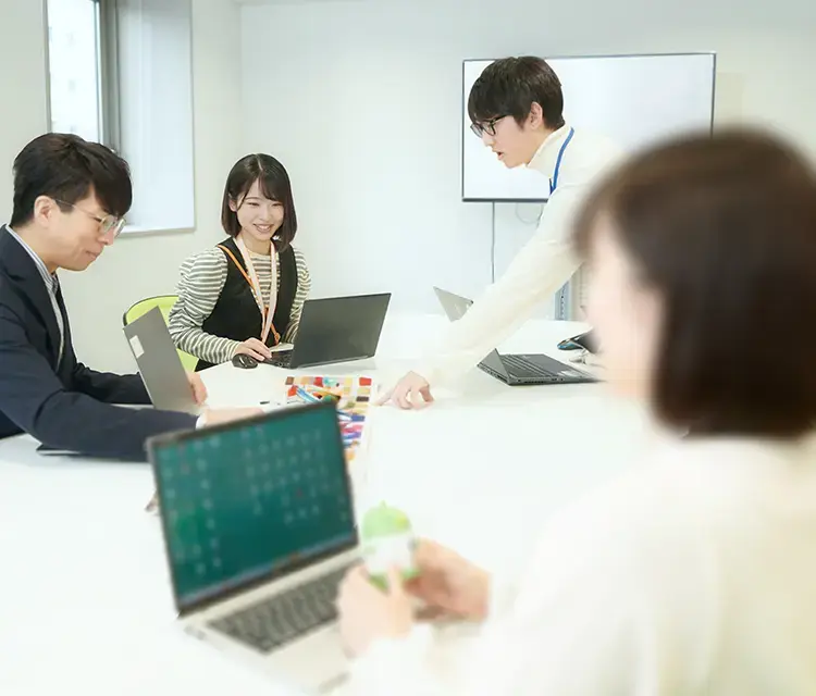 写真：会議テーブルで各自ノートPCを扱いながら打ち合わせをしている男性社員2名と女性社員2名